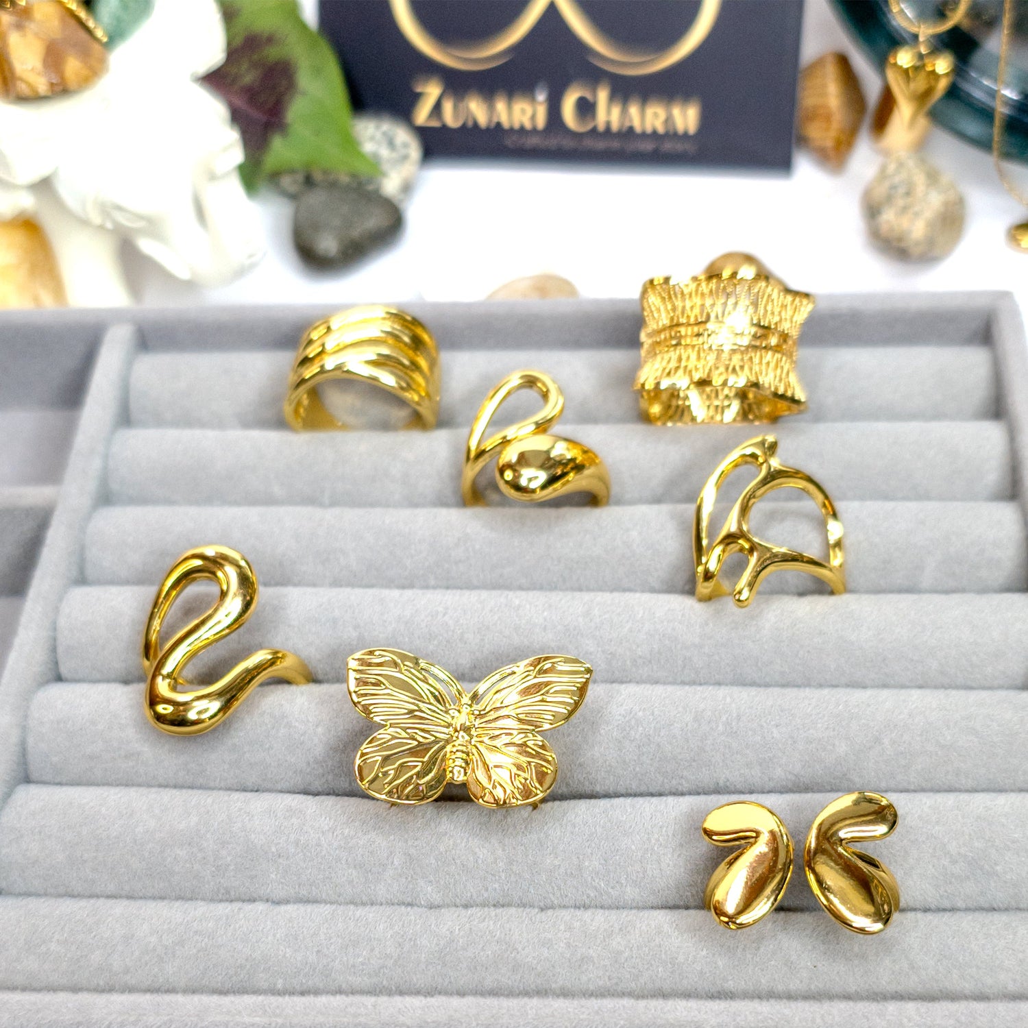 Collection of gold statement rings from Zunari Charm displayed on a grey velvet jewelry tray, featuring designs like butterfly ring, molten drop ring, infinity flow ring, bow knot, and abstract sculptural styles.