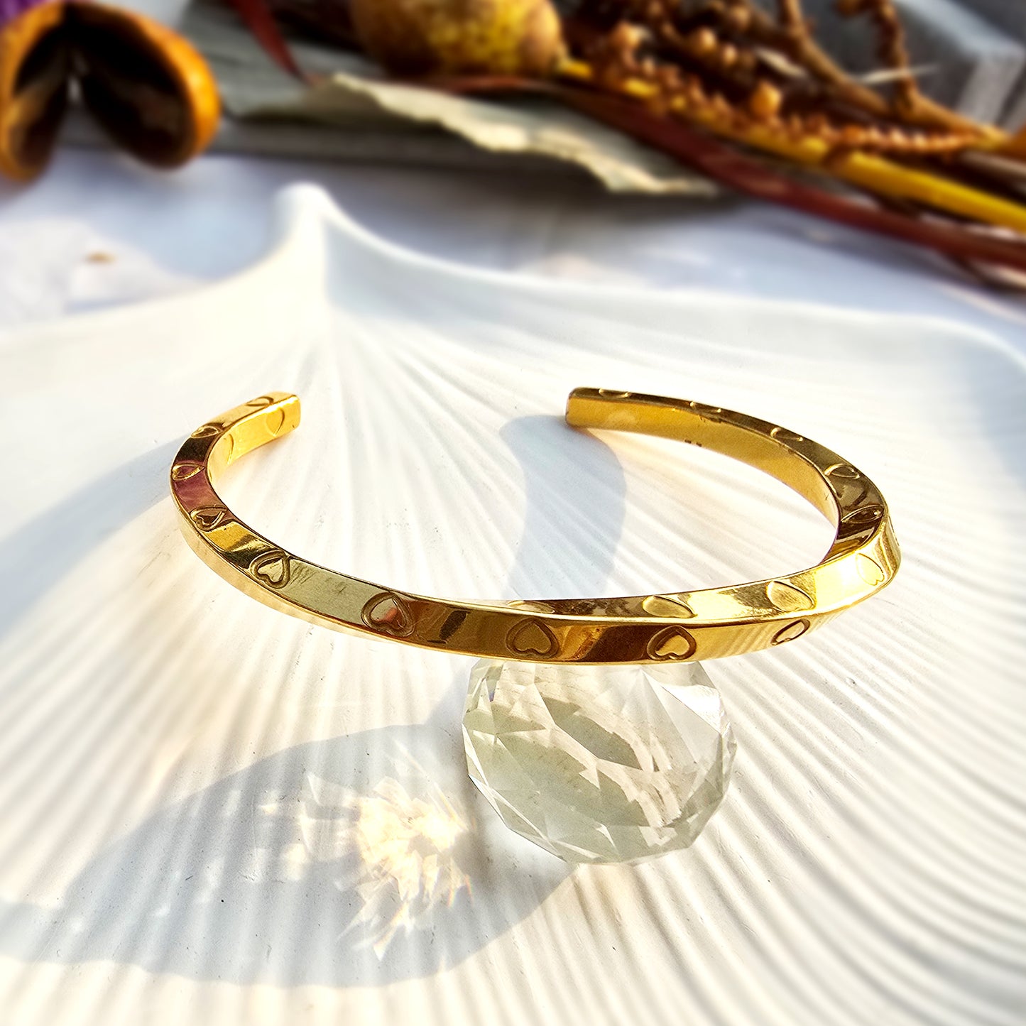 Gold twisted ribbon cuff bracelet with engraved hearts resting on a white shell.