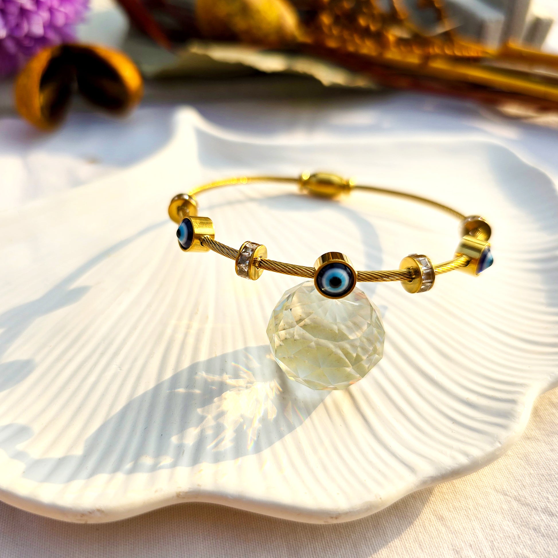 Luxury gold cable bangle with protective eye charms styled on a white shell tray.