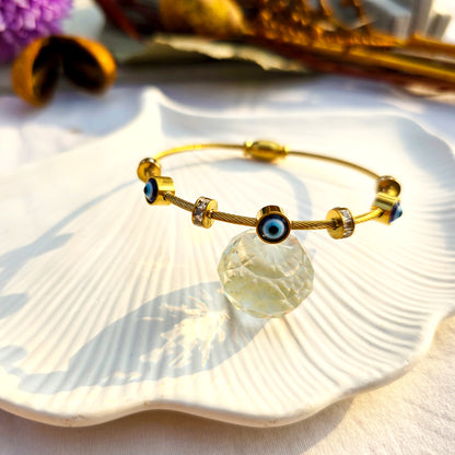Luxury gold cable bangle with protective eye charms styled on a white shell tray.