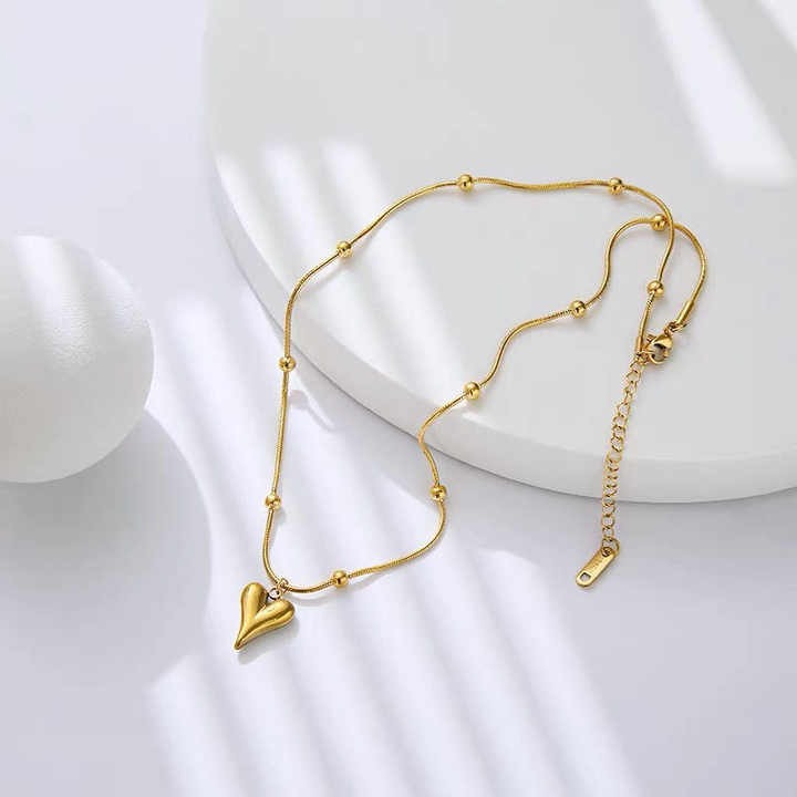 Gold-toned teardrop heart necklace with beaded chain laid flat on white minimal background