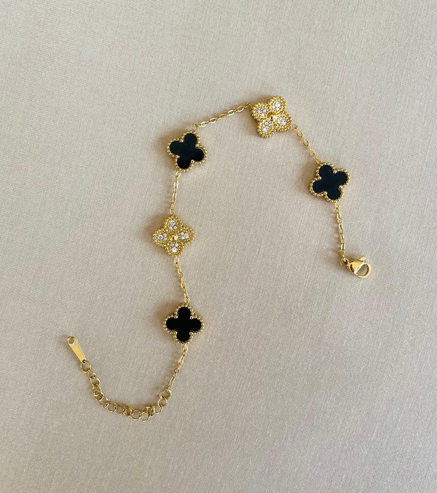 Gold bracelet with alternating black clover and crystal charms laid flat on neutral background