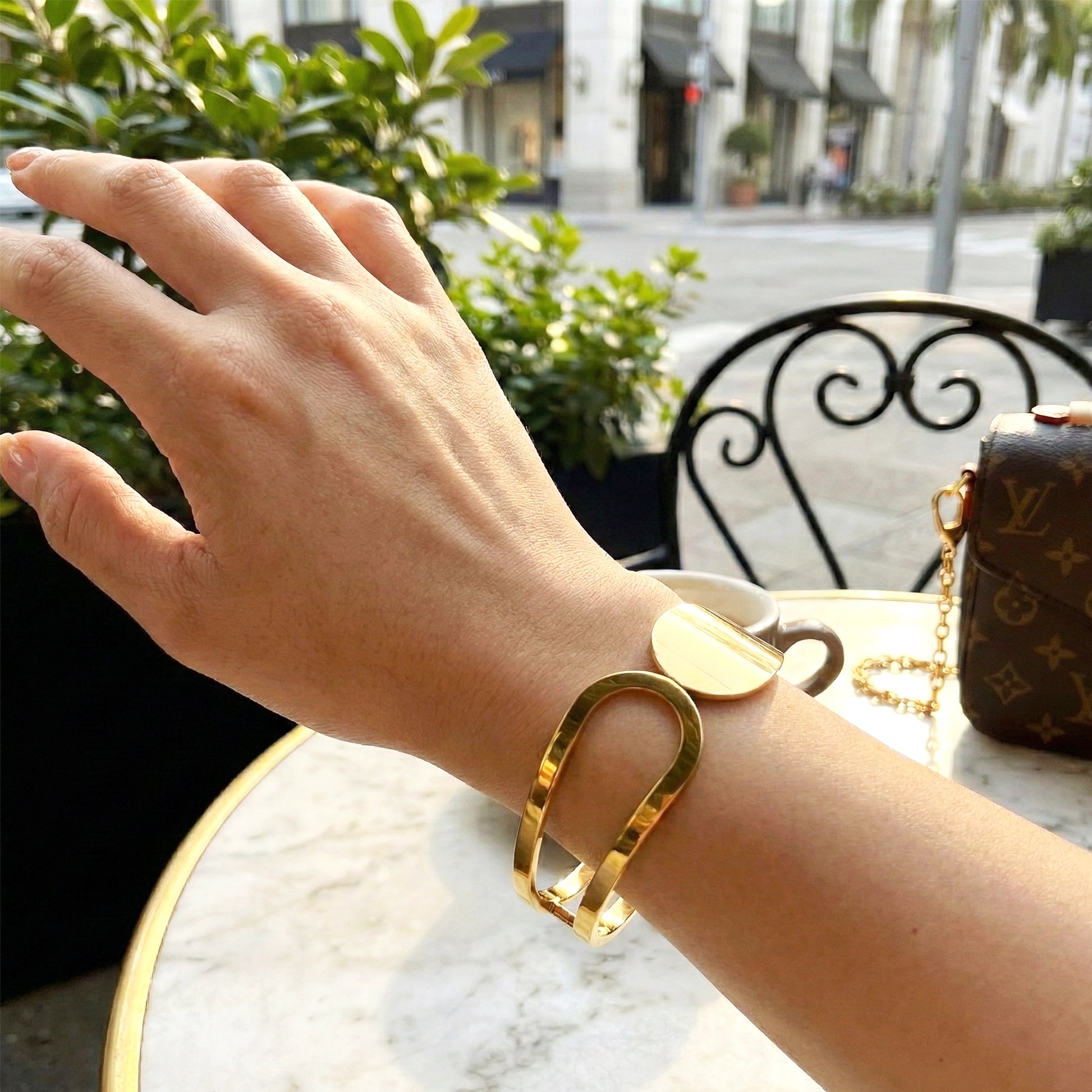 Woman wearing bold gold arc bangle styled with luxury handbag at a cafe.