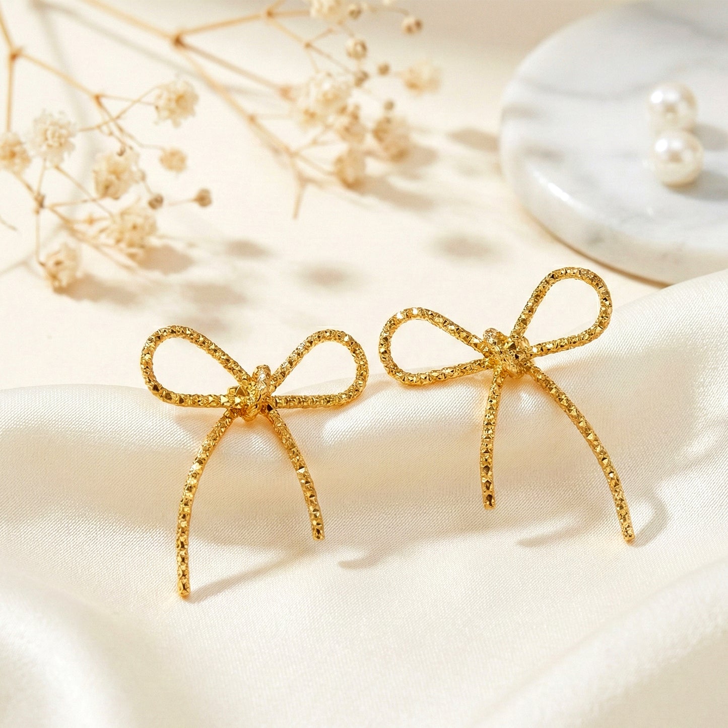Aesthetic flat lay of textured gold bow earrings on white silk fabric with dried flowers background.
