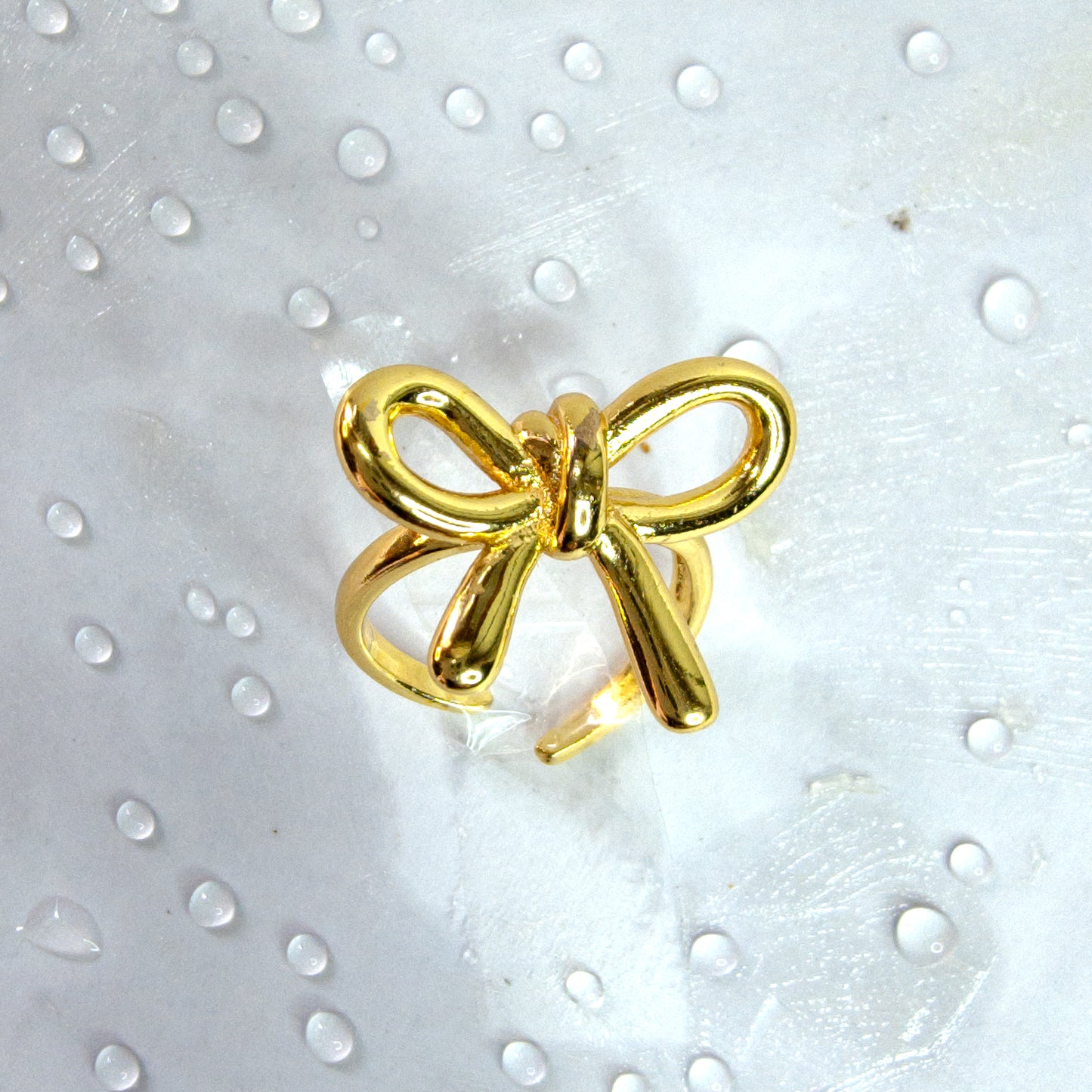 Close-up of gold bow knot adjustable ring with glossy ribbon design, styled on a white surface with water droplets – Zunari Charm jewelry.