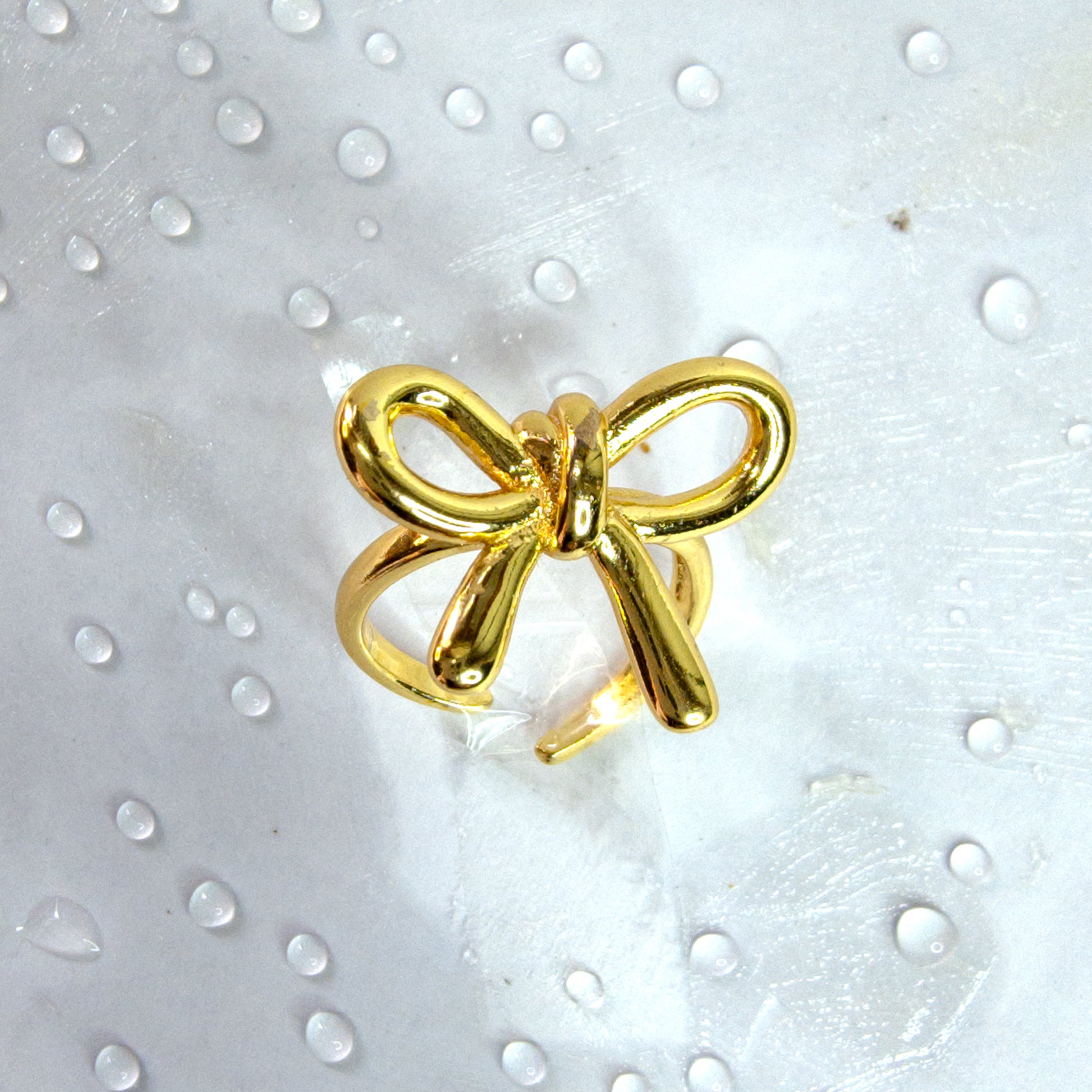 Close-up of gold bow knot adjustable ring with glossy ribbon design, styled on a white surface with water droplets – Zunari Charm jewelry.