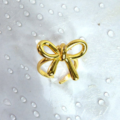 Close-up of gold bow knot adjustable ring with glossy ribbon design, styled on a white surface with water droplets – Zunari Charm jewelry.