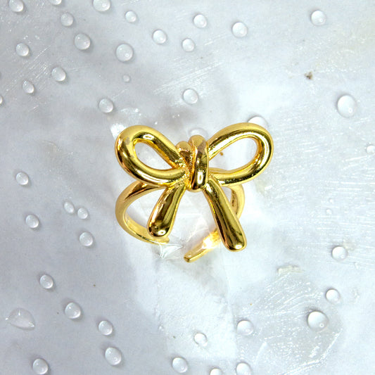 Close-up of gold bow knot adjustable ring with glossy ribbon design, styled on a white surface with water droplets – Zunari Charm jewelry.