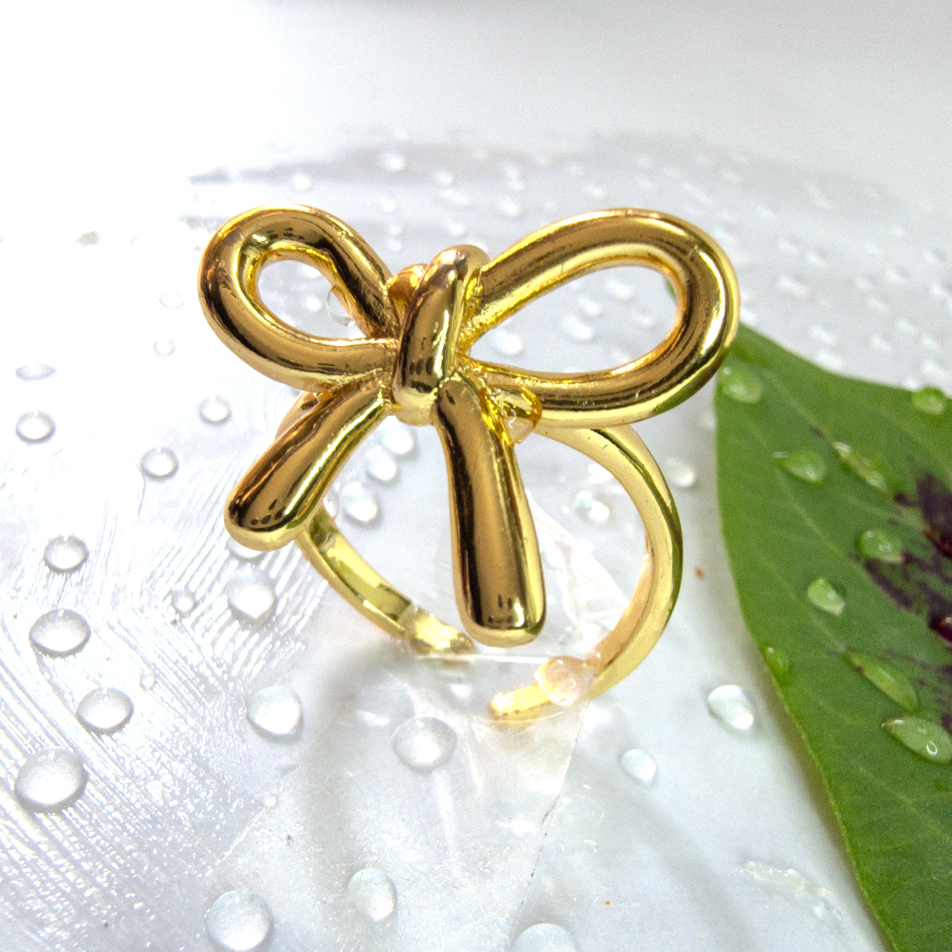 Gold bow knot ring with polished ribbon design, placed on a white background with water droplets and green leaf accent – Zunari Charm accessory.