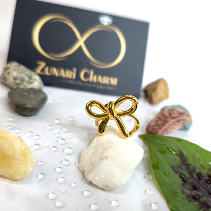 Gold bow knot statement ring with a delicate ribbon design, displayed on a white stone with Zunari Charm branding in background.