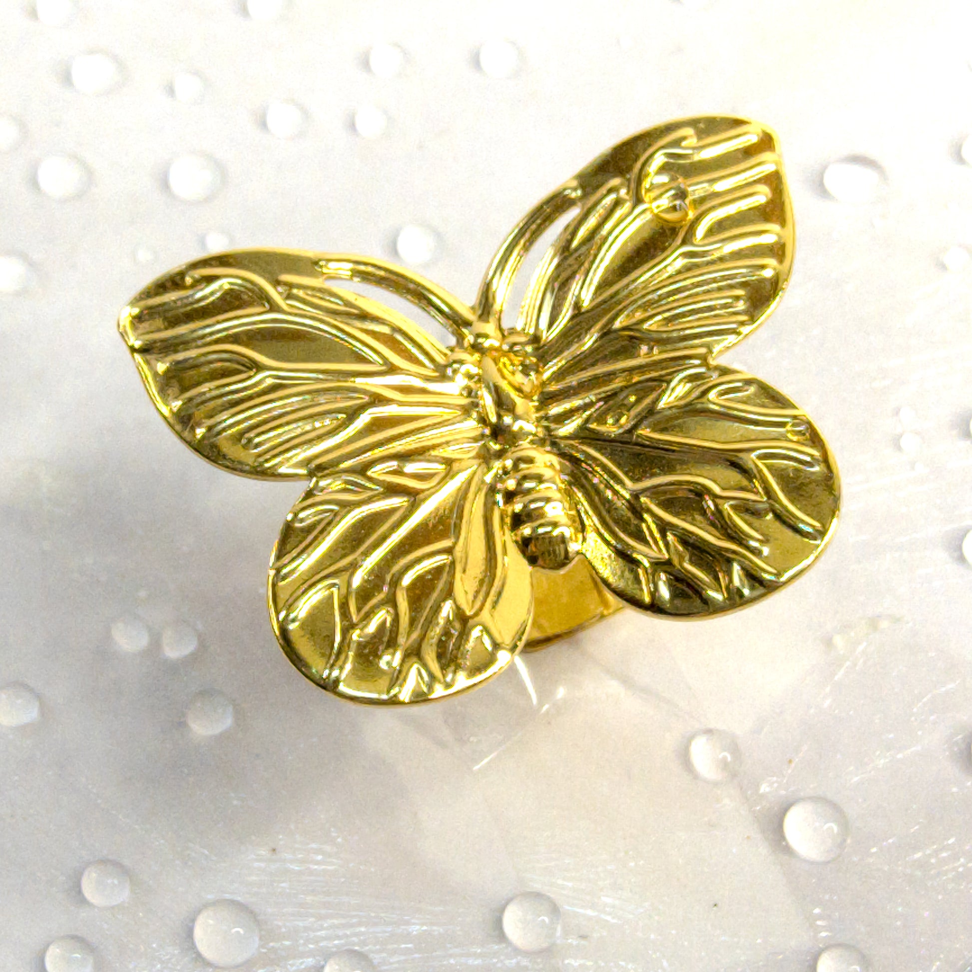 Gold butterfly adjustable ring with intricate detailing, styled flat on a white surface with droplets – Zunari Charm statement accessory.