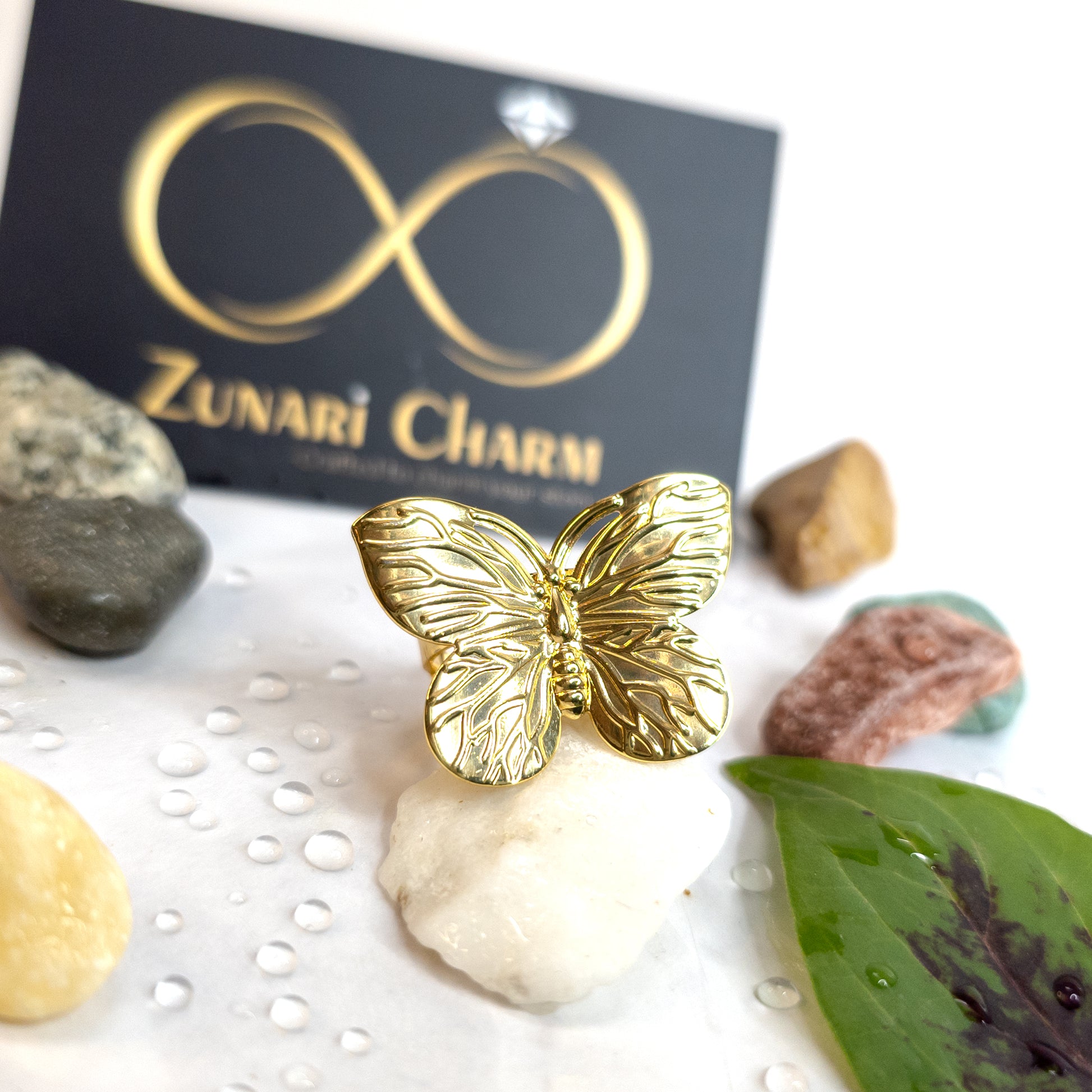 Gold butterfly statement ring with textured wing detailing, displayed on a white stone with water droplets, Zunari Charm jewelry branding in the background.