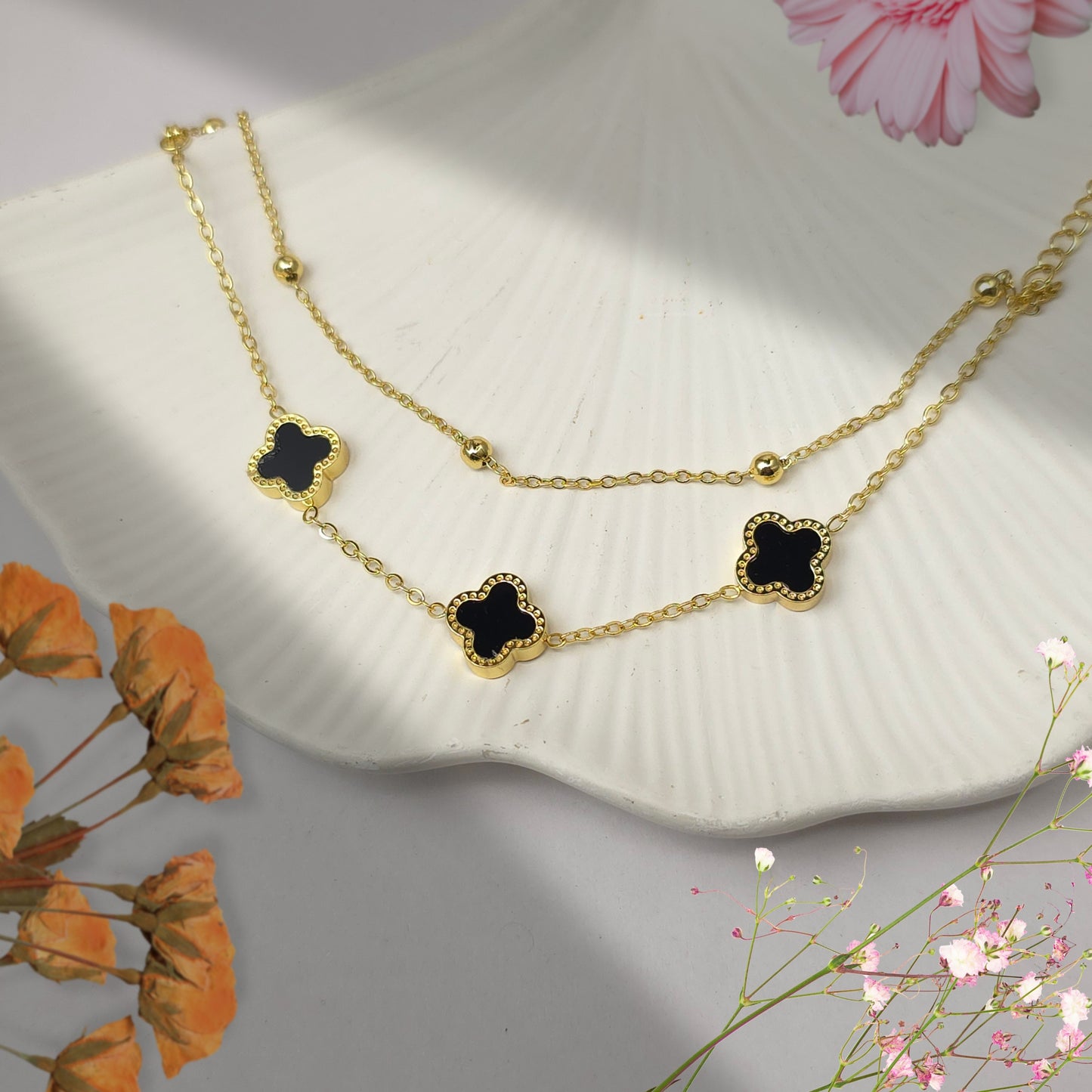 Black clover layered gold bracelet displayed on white shell with floral aesthetic background minimal luxury jewelry
