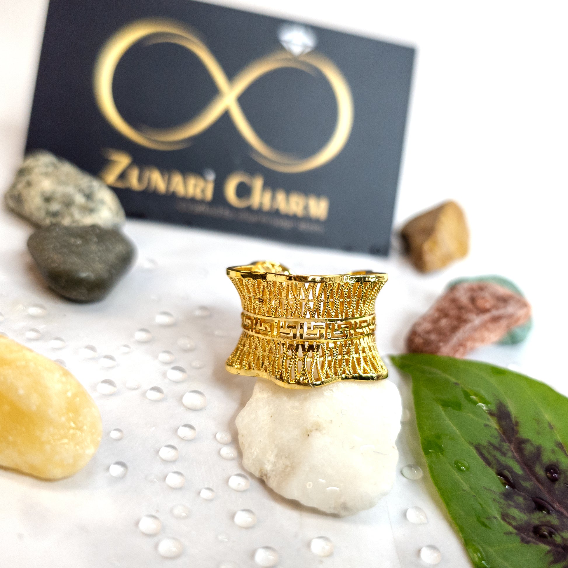 Gold ring on a rock with 'Zunari Charm' branding in the background. Gold-toned Empress Mesh Ring displayed on white stone with water droplets, highlighting its wide band texture.