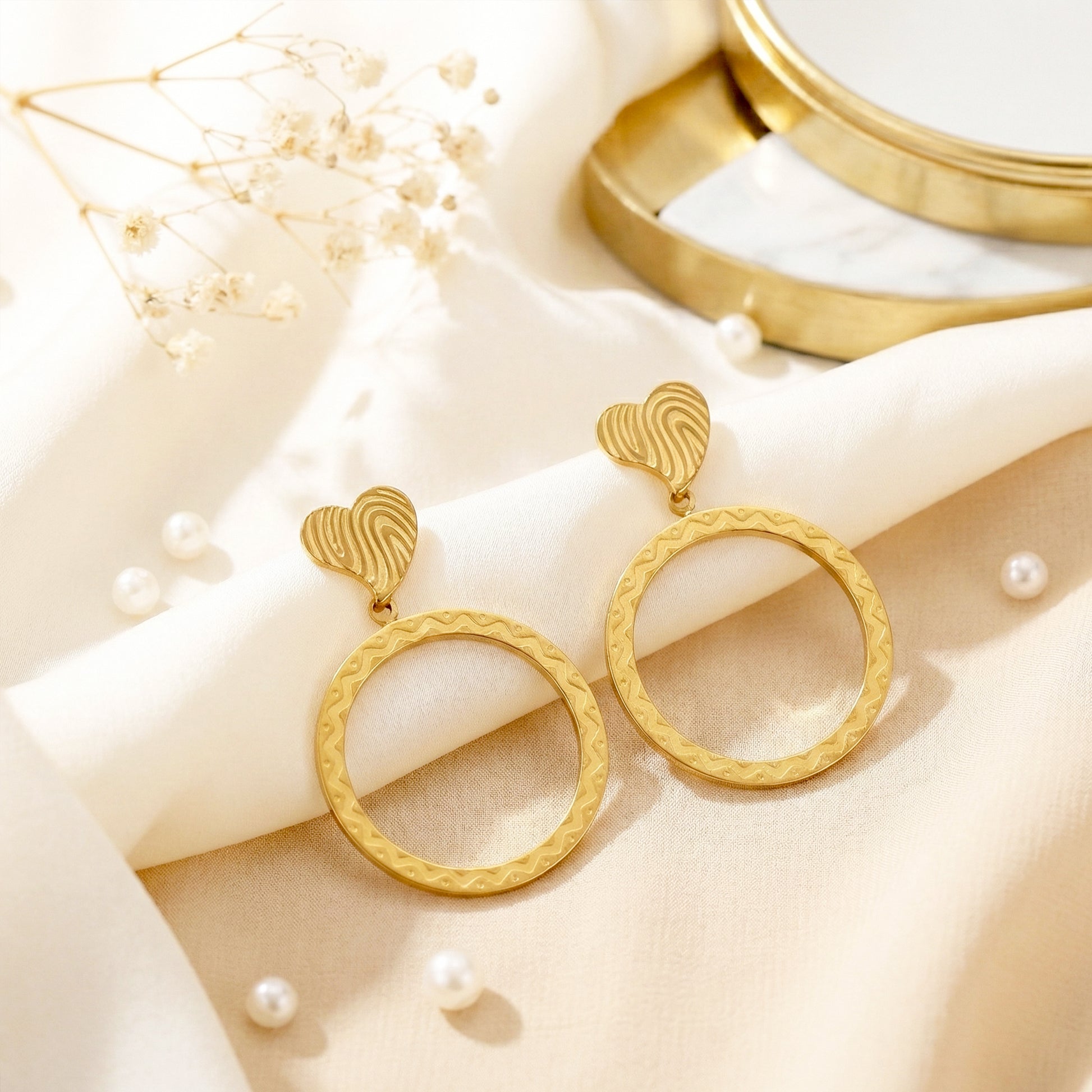 Aesthetic photography of gold etched heart earrings on cream silk fabric with white pearls and dried flowers.