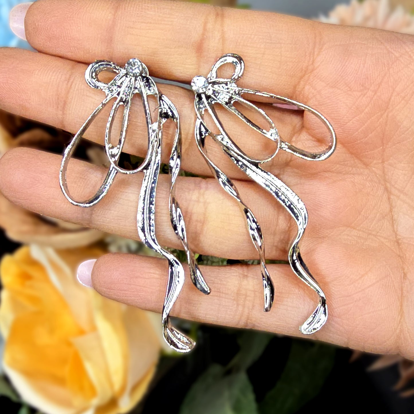 Close-up of silver-toned bow earrings held in a hand to show the sparkling crystal knot and reflective metallic finish.