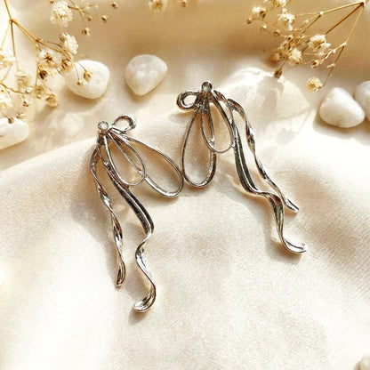 Aesthetic photography of oversized silver bow earrings on a cream silk cloth with white decorative stones and dried flowers.