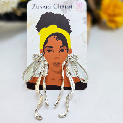 Exaggerated silver bow dangles presented on a Zunari Charm branded card featuring an illustrated woman in a yellow turban.