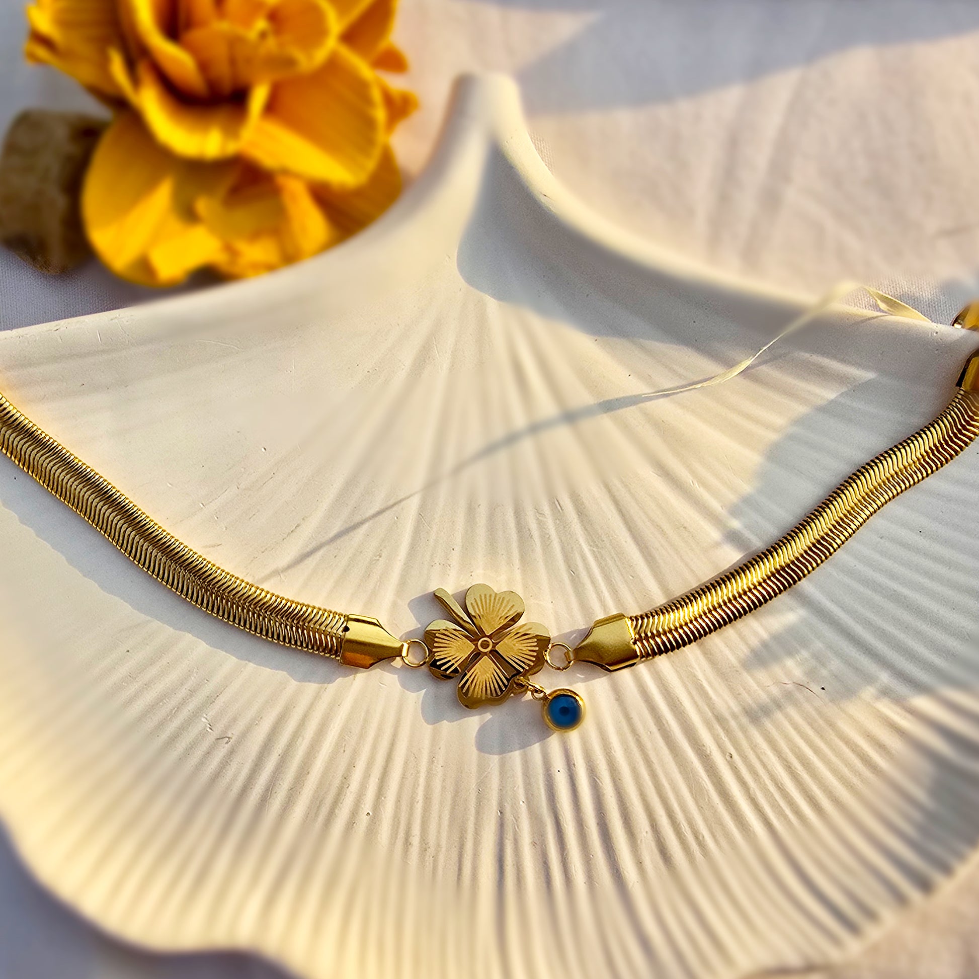 Gold snake chain bracelet with four-leaf clover and blue evil eye charm on white shell tray.