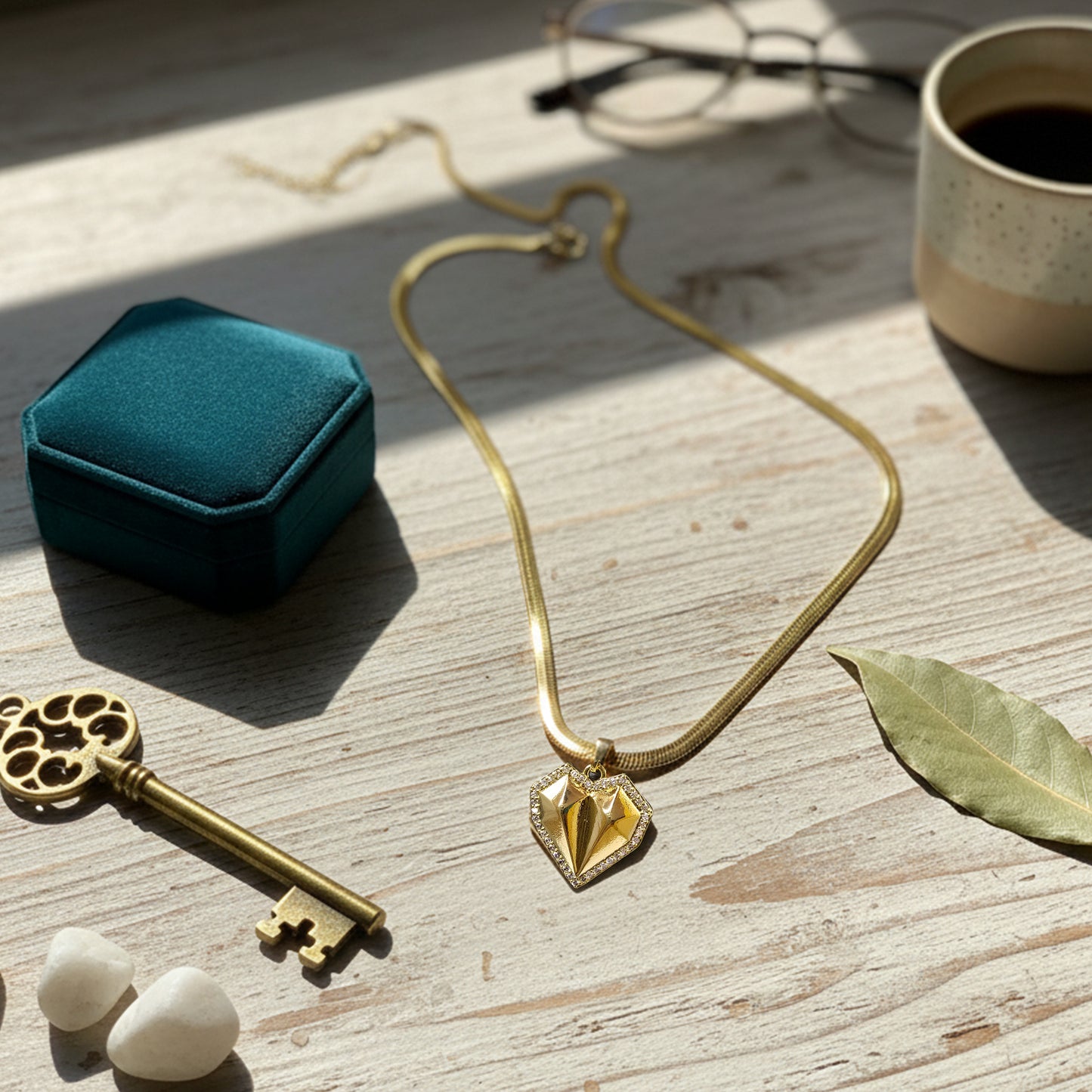 Geometric heart necklace with herringbone chain styled on a light wooden table with coffee and vintage key.