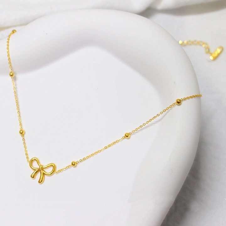 Gold-toned minimalist bow necklace styled on a curved ceramic surface for a soft, luxurious visual appeal.