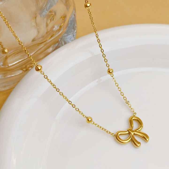 Flat lay of the gold-toned bow necklace placed on a ceramic dish with a clear glass in the background.