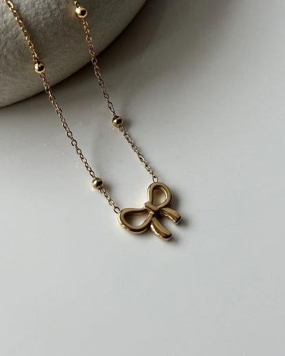 Close-up of a delicate gold-toned necklace featuring a minimalist bow pendant and small gold beads, styled on a neutral background.