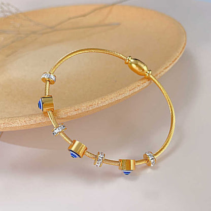 Top view of gold twisted-wire bracelet with crystal rings and blue evil eye barrel beads, placed on a beige dish.