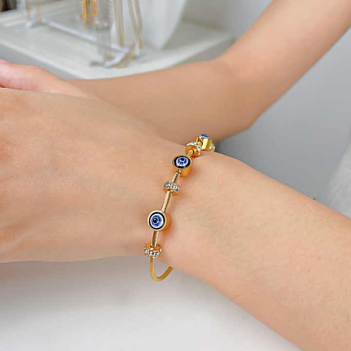 Model wearing a gold-tone evil eye bangle adorned with rhinestones and blue enamel evil eye beads.