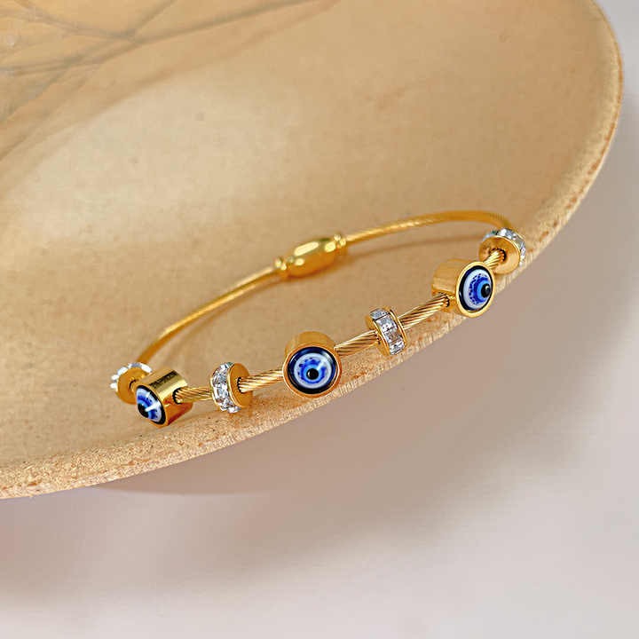 Close-up of a golden cable-style evil eye bangle with crystal and blue enamel charms, placed on a peach-toned dish.