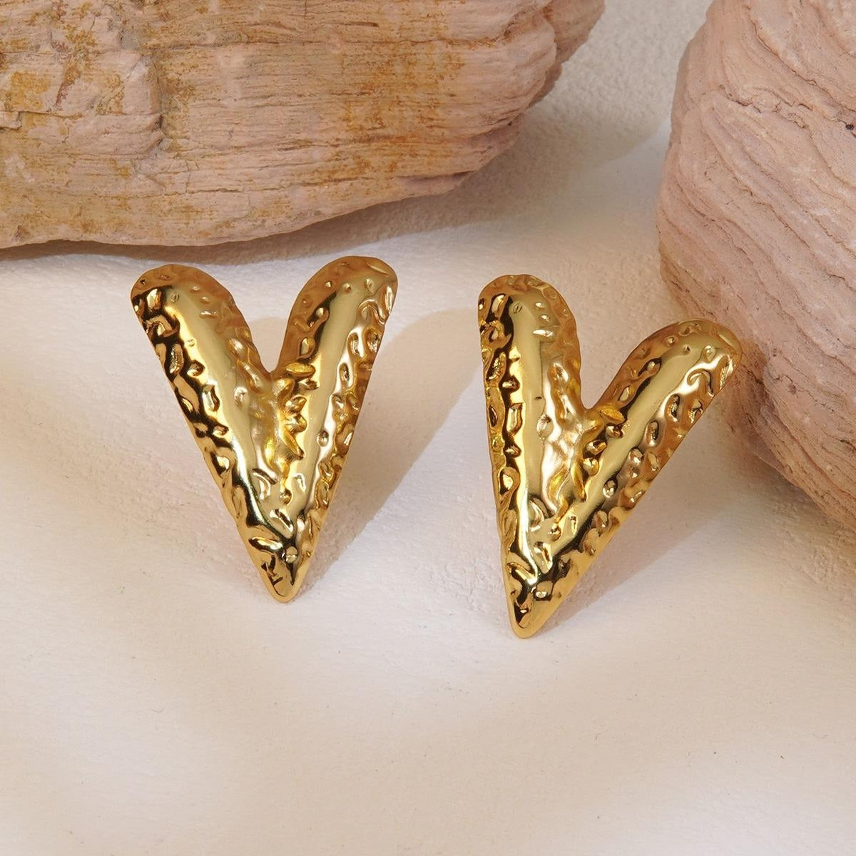 Textured V-shaped earrings with a reflective hammered gold finish set between natural stone props for an earthy yet bold display
