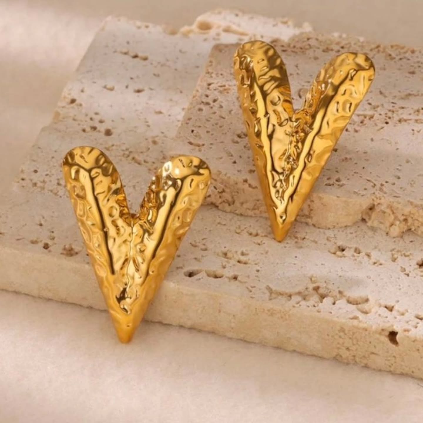 Hammered gold V-shaped earrings placed on a neutral stone platform, showcasing their edgy and artistic surface design