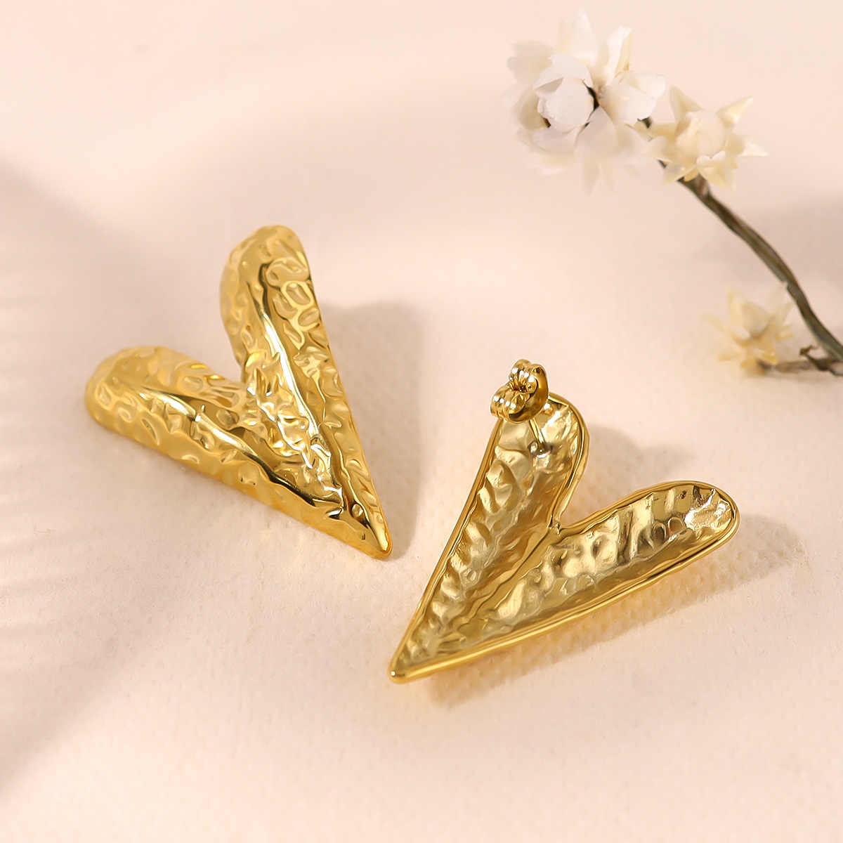 Bold, hammered gold-toned V-shaped earrings with sculptural texture placed on a soft pastel fabric background beside delicate white flowers.