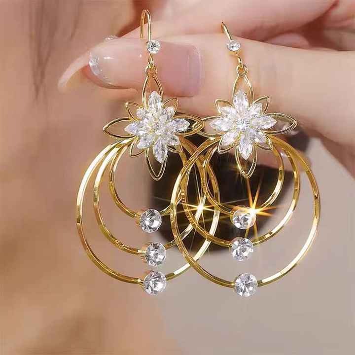 Gold earrings with floral designs held by a hand against a neutral background