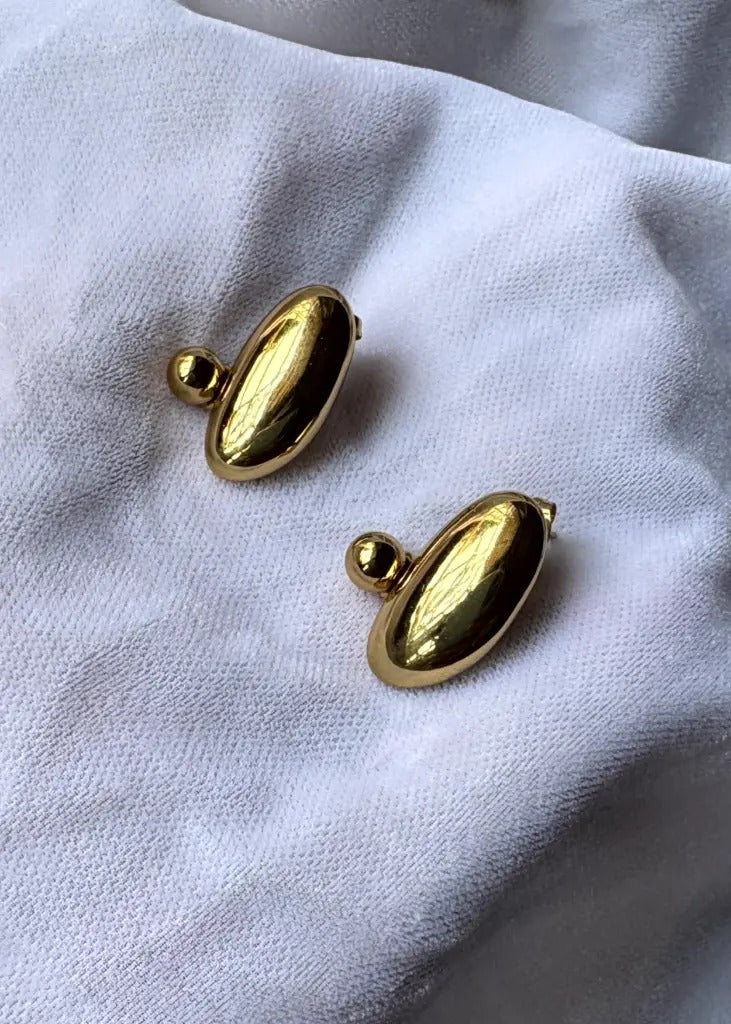 Pebble-shaped gold earrings placed on a soft white fabric background