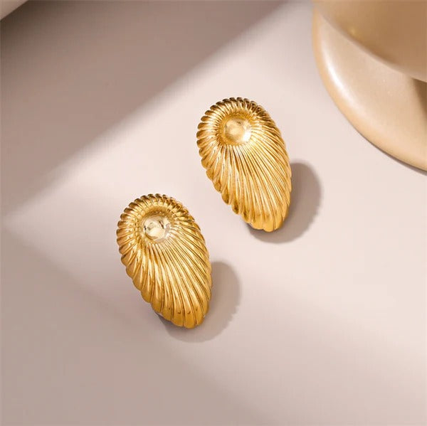 Pair of golden shell-inspired earrings displayed under soft lighting on a neutral background, highlighting the ridged detailing and elegant shine