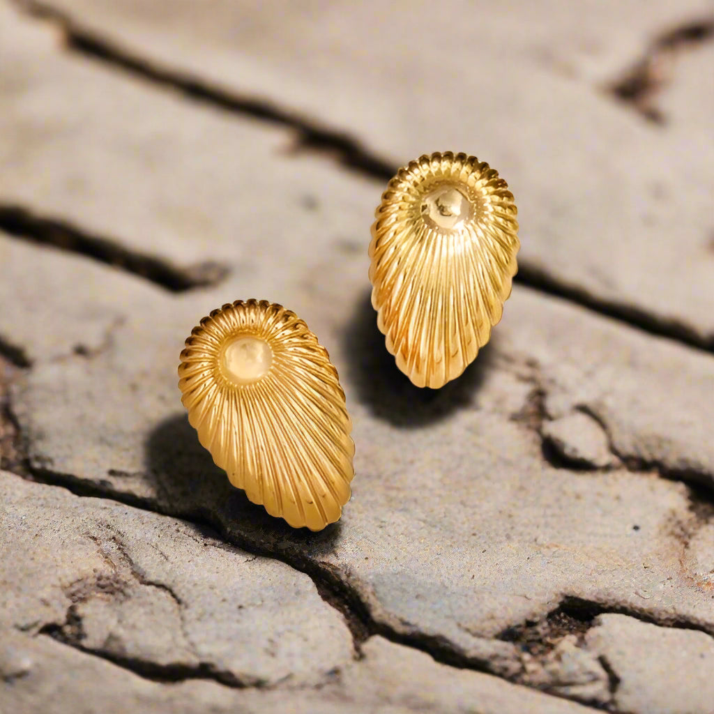 Top view of golden shell-shaped earrings showcasing their spiral ridges and high-shine metallic texture on cobblestone bac
ground