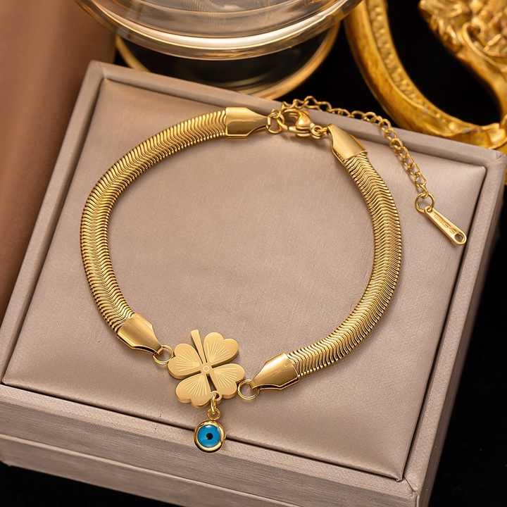 Gold snake chain bracelet with clover and evil eye charms placed on beige box