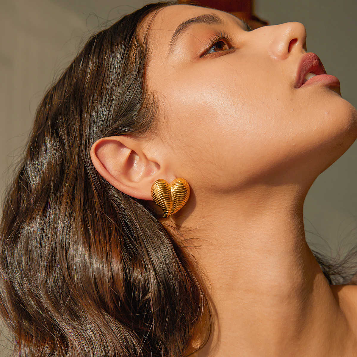 Model wearing bold gold shell-textured heart earrings, styled with minimal makeup and side-parted hair