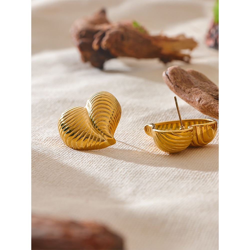 Heart-shaped gold shell earrings resting on soft cream fabric with earthy wood props, showcasing front and back