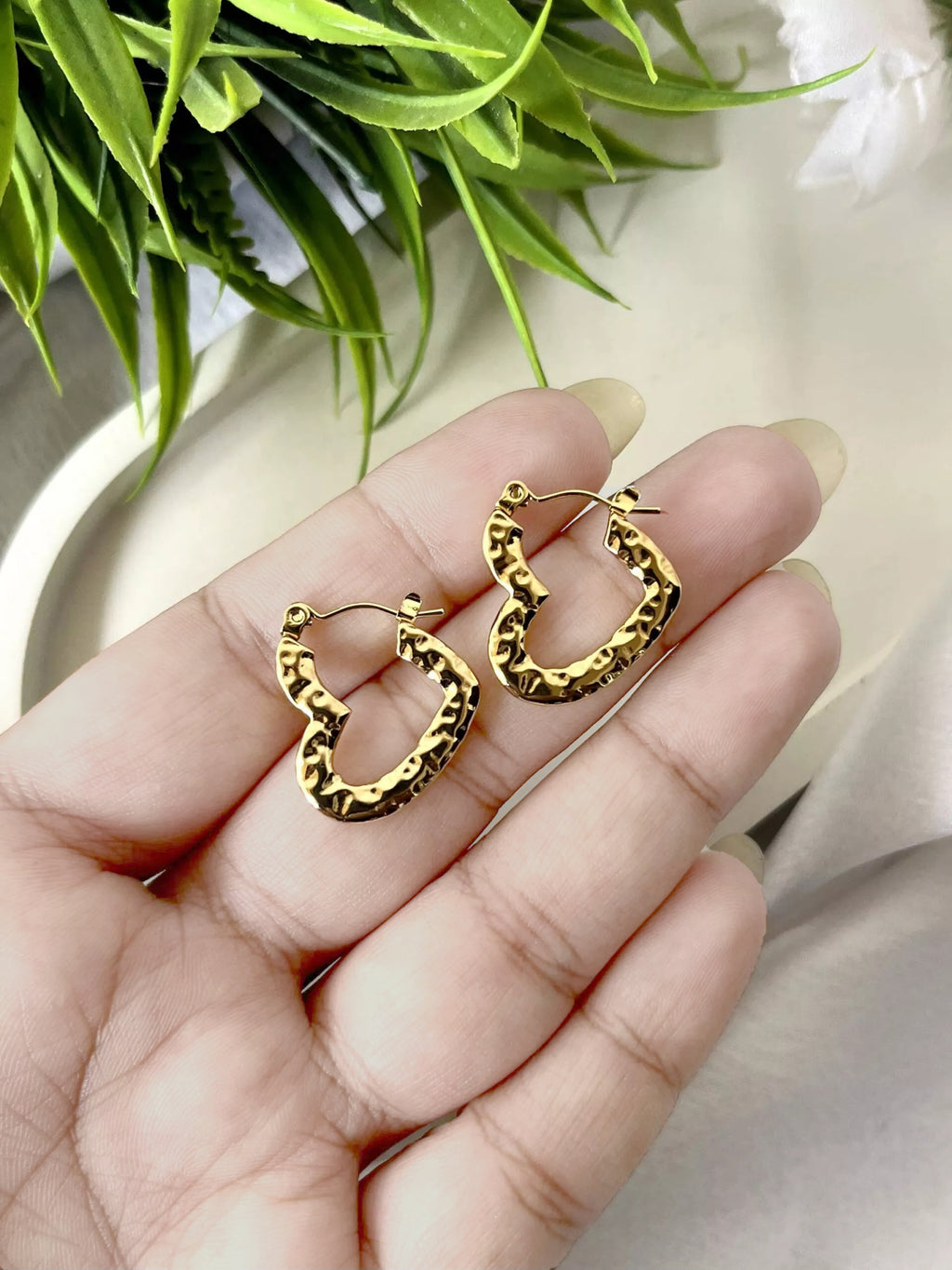 Hand holding textured gold heart earrings with green leaves and white flowers in the background