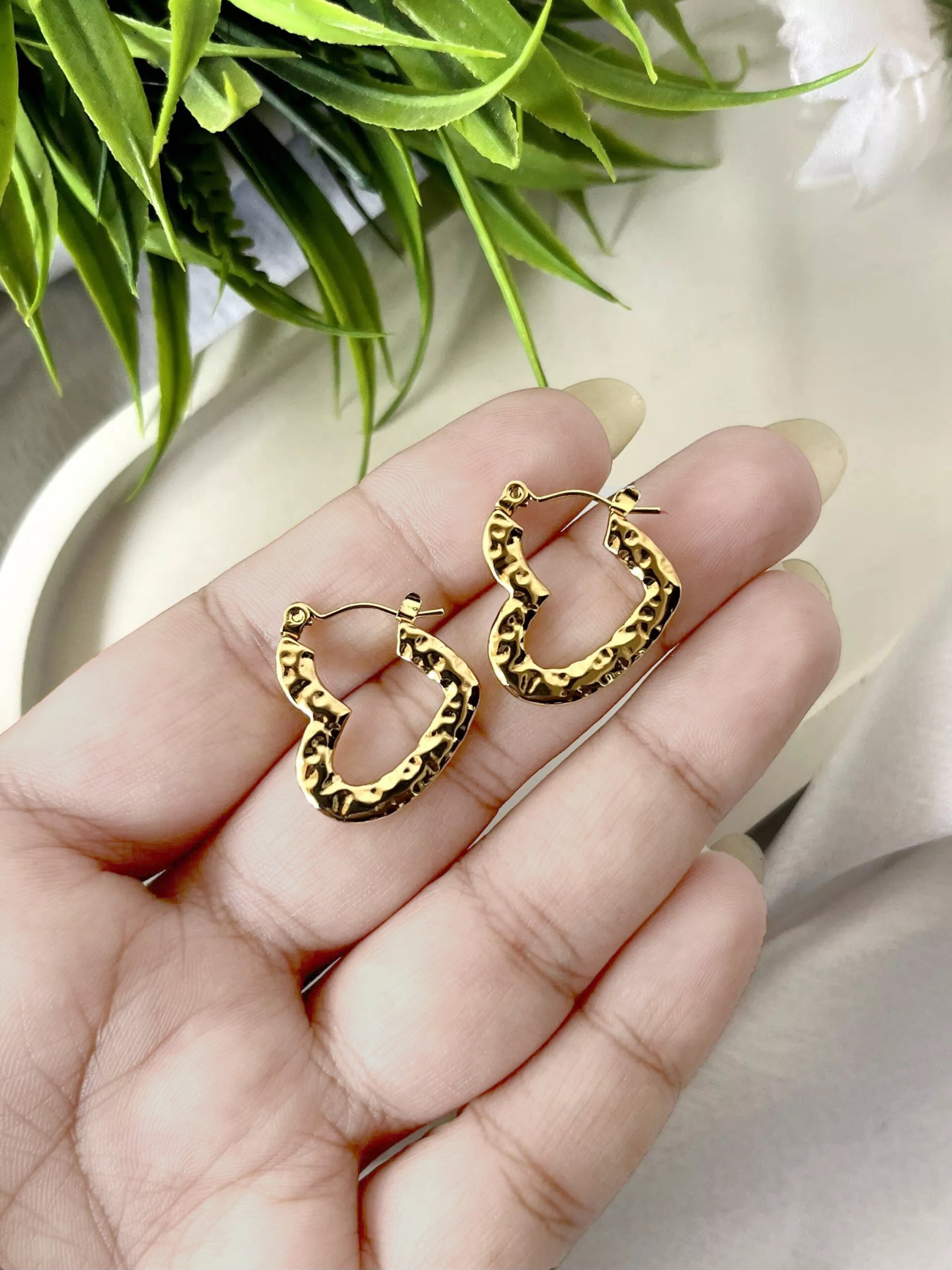 Hand holding textured gold heart earrings with green leaves and white flowers in the background