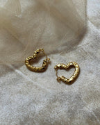 Textured heart-shaped gold hoop earrings resting on soft beige mesh fabric