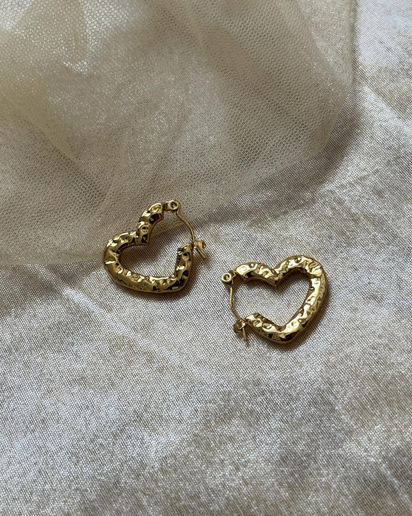 Textured heart-shaped gold hoop earrings resting on soft beige mesh fabric