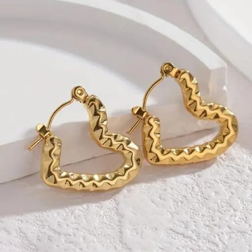 Close-up of textured gold heart hoop earrings placed on a minimal white backdrop with curved props