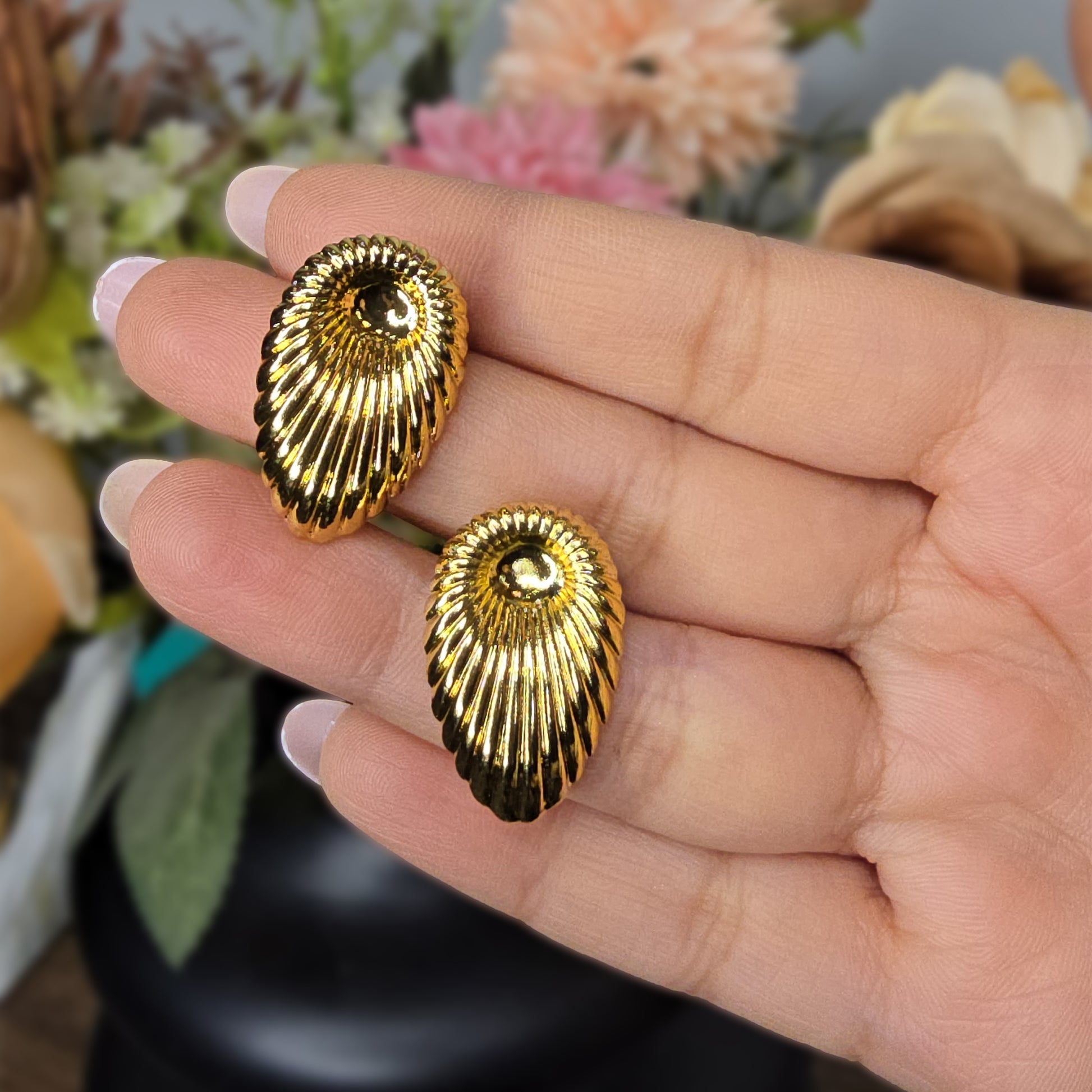 A hand holding two large gold fluted shell earrings to show the reflective shine and scale of the pieces.