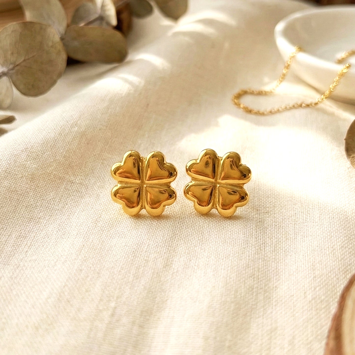 Aesthetic photography of gold clover studs on a cream linen cloth with a soft sunlit background.