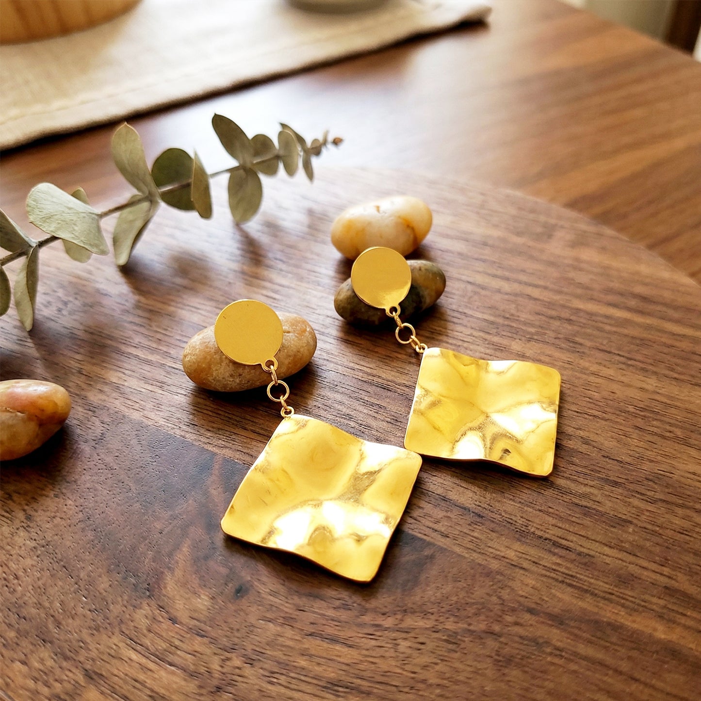 Aesthetic flat lay of liquid gold earrings on a wooden board with smooth river stones and a sprig of eucalyptus.