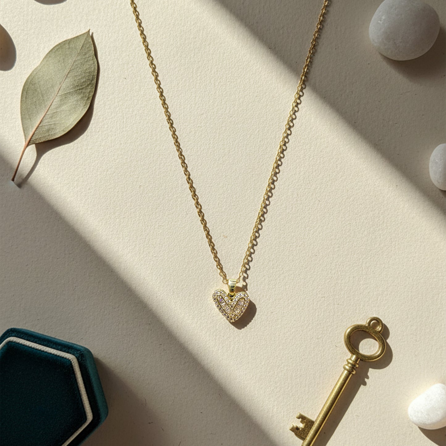 Heart necklace styled with a velvet box, eucalyptus leaf, and key on a neutral background in bright sunlight.