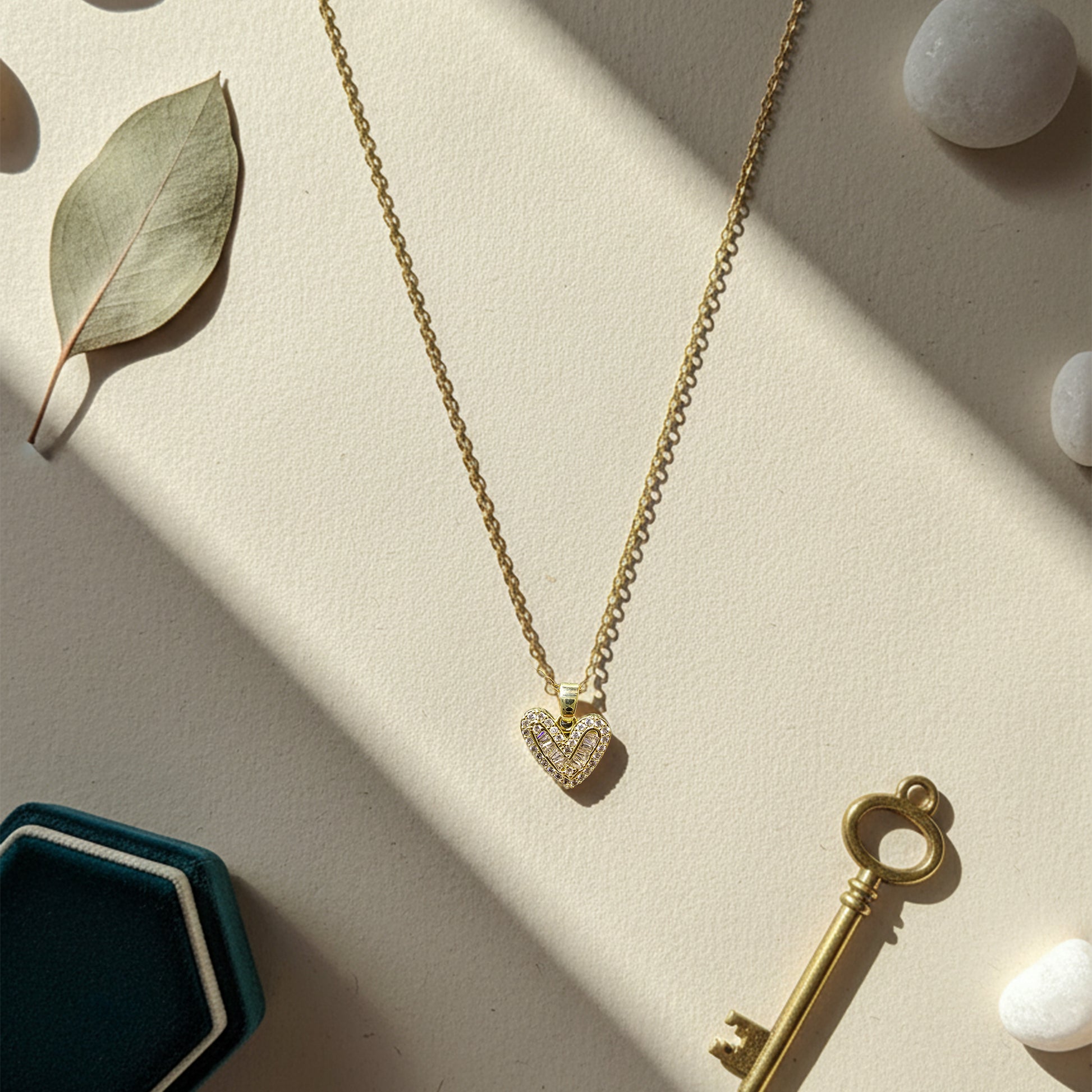Heart necklace styled with a velvet box, eucalyptus leaf, and key on a neutral background in bright sunlight.